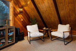 Una habitación acogedora con paredes de madera, una estantería, dos sillones blancos con cojines, una mesita y una lámpara.