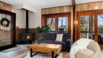 A cozy living room with a fireplace, a sofa, a coffee table, and a large window with a view of trees.