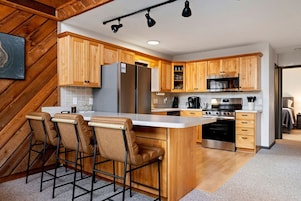Una cocina moderna con armarios de madera, un refrigerador de acero inoxidable, un microondas y un horno empotrado. Hay taburetes en la isla y un cuadro en la pared.