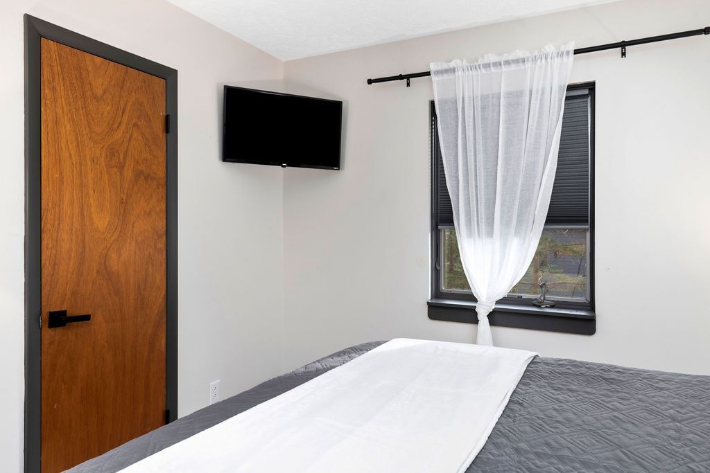 Ein Schlafzimmer mit einem Bett, einer Holztür, einem Fernseher und einem Fenster mit Vorhang.