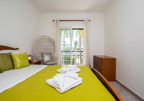 A hotel room with a bed, a wooden dresser, a round wicker chair, and a view of greenery through a window. - Casa das Chaminés – In Conceicao de Tavira (Tavira)