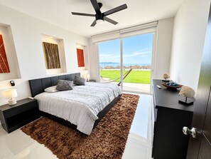 Un dormitorio moderno con una cama grande, un ventilador de techo y vistas a un paisaje verde a través del ventanal.