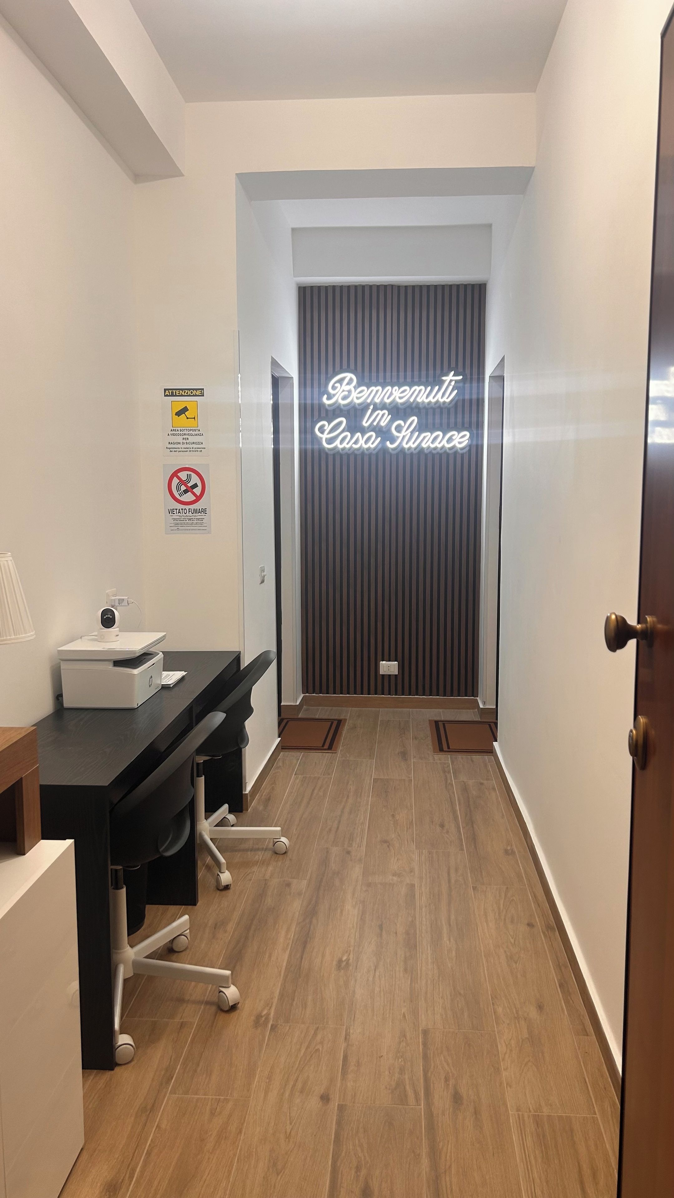 Un pasillo con mostrador de recepción, un letrero de neón que dice 'Welcome to Casa Sirace' y un cartel de advertencia de seguridad.