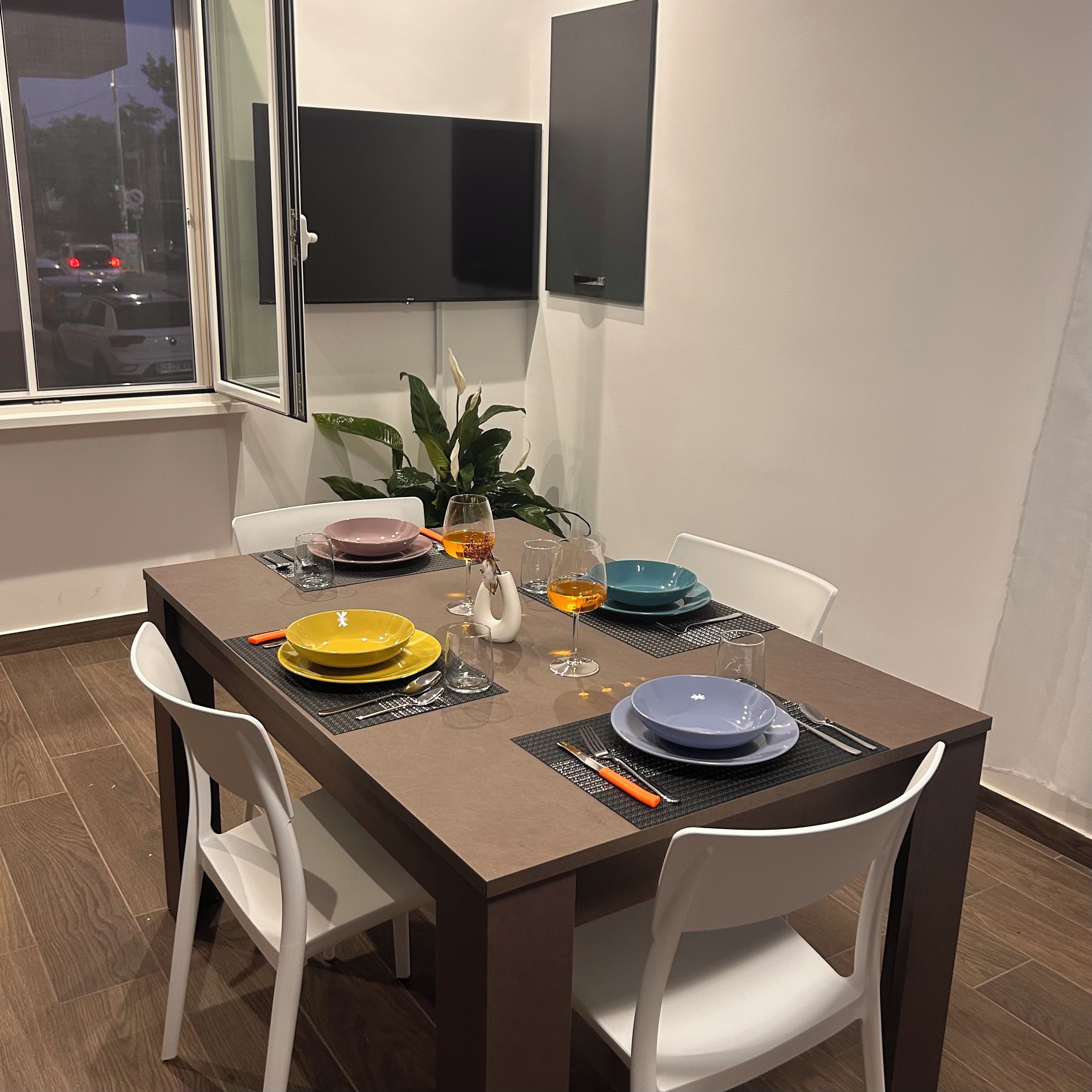 Un comedor con una mesa dispuesta para dos personas, sillas blancas y una planta en maceta.