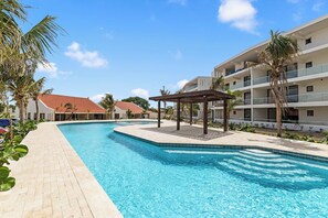 A swimming pool with a pergola, palm trees, and a building with a red roof. - J&M Coral - Modern 2BR & Shared Pool near Mambo (Willemstad)