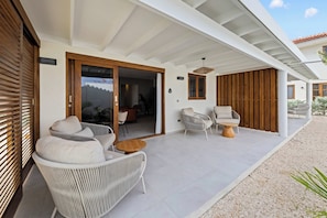A modern outdoor patio with wicker furniture, a wooden table, and a view of the interior. - J&M Coral - Modern 2BR & Shared Pool near Mambo (Willemstad)
