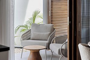A modern outdoor seating area with a grey upholstered chair and a round wooden table, set against a backdrop of a wooden shutter and a palm tree. - J&M Coral - Modern 2BR & Shared Pool near Mambo (Willemstad)