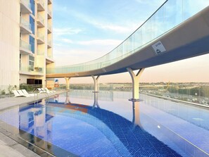 Una piscina a sfioro moderna con un ponte in vetro, sedie a sdraio e vista sulla città.