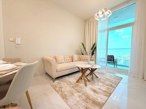 A modern living room with a sofa, a dining table, and a view of the city through large windows. - Stylish 1BR Apartment - Skyz by Danube - Arjan (Dubai)
