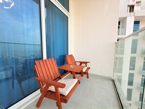 Un balcone con due sedie di legno, un tavolino, una ringhiera in vetro e vista sulla città attraverso ampie finestre.