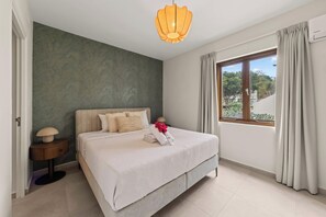A bedroom with a bed, a nightstand, a lamp, a window with curtains, and a view of greenery outside. - J&M Trupial | Stylish Caribbean Stay in Vredenberg (Willemstad)