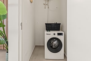 A compact laundry room with a white washing machine, a small black basket, and a potted plant. - J&M Trupial | Stylish Caribbean Stay in Vredenberg (Willemstad)