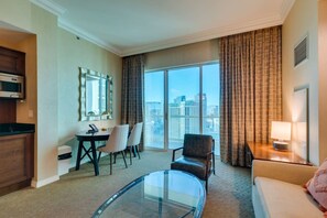Quarto de hotel com área de refeições, sofá, mesa de centro de vidro e vista para a paisagem urbana.