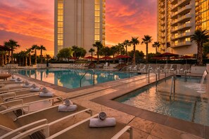 Ein Hotelpoolbereich mit Liegestühlen und Sonnenschirmen, umgeben von Palmen und Hochhäusern.