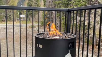 A black outdoor fire pit on a wooden deck with a metal railing, surrounded by trees and a mountainous backdrop.