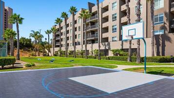 A basketball court with a net, surrounded by palm trees and a multi-story building with balconies.