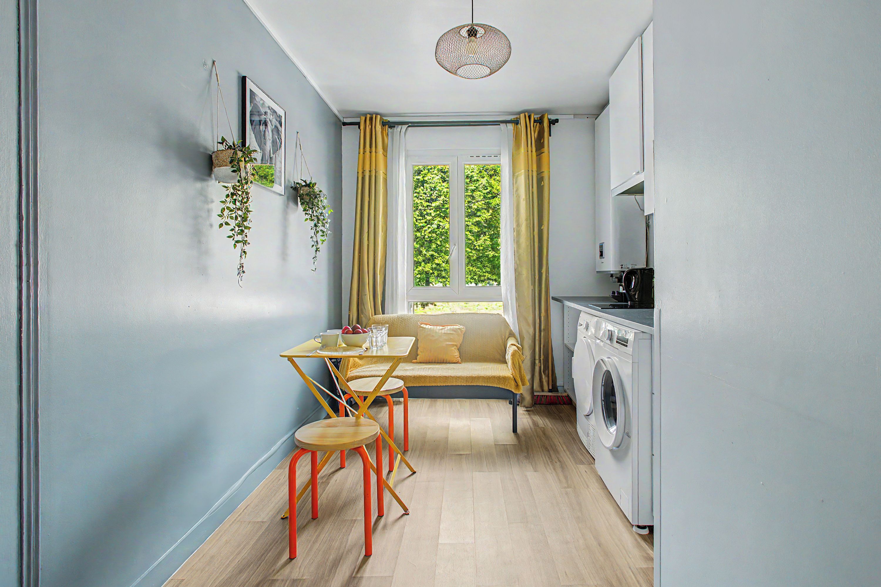 A compact kitchen with a washing machine, a dining area with a sofa and table, and a window with curtains.