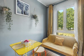 Ein Zimmer mit einem gelben Tisch, einem Sofa, einem Fenster mit Vorhängen und zwei hängenden Pflanzen.