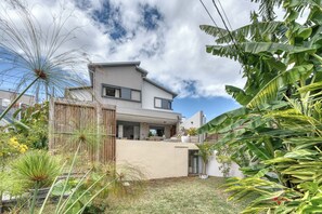 Ein modernes, zweistöckiges Haus mit flachem Dach, einem Balkon und einem gepflegten Garten mit verschiedenen Pflanzen und einer Rasenfläche.