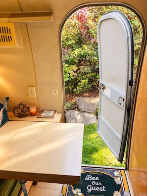 A cozy RV interior with a bed, a small table with a book and candle, and a door leading to a garden. - Vintage Airstream Caravel Retreat Near Beach & Sea-Tac (Des Moines)