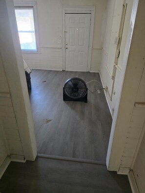 Ein Zimmer mit weißer Tür, einem Fenster mit Jalousien, einem Deckenventilator und einer sichtbaren Steckdose.