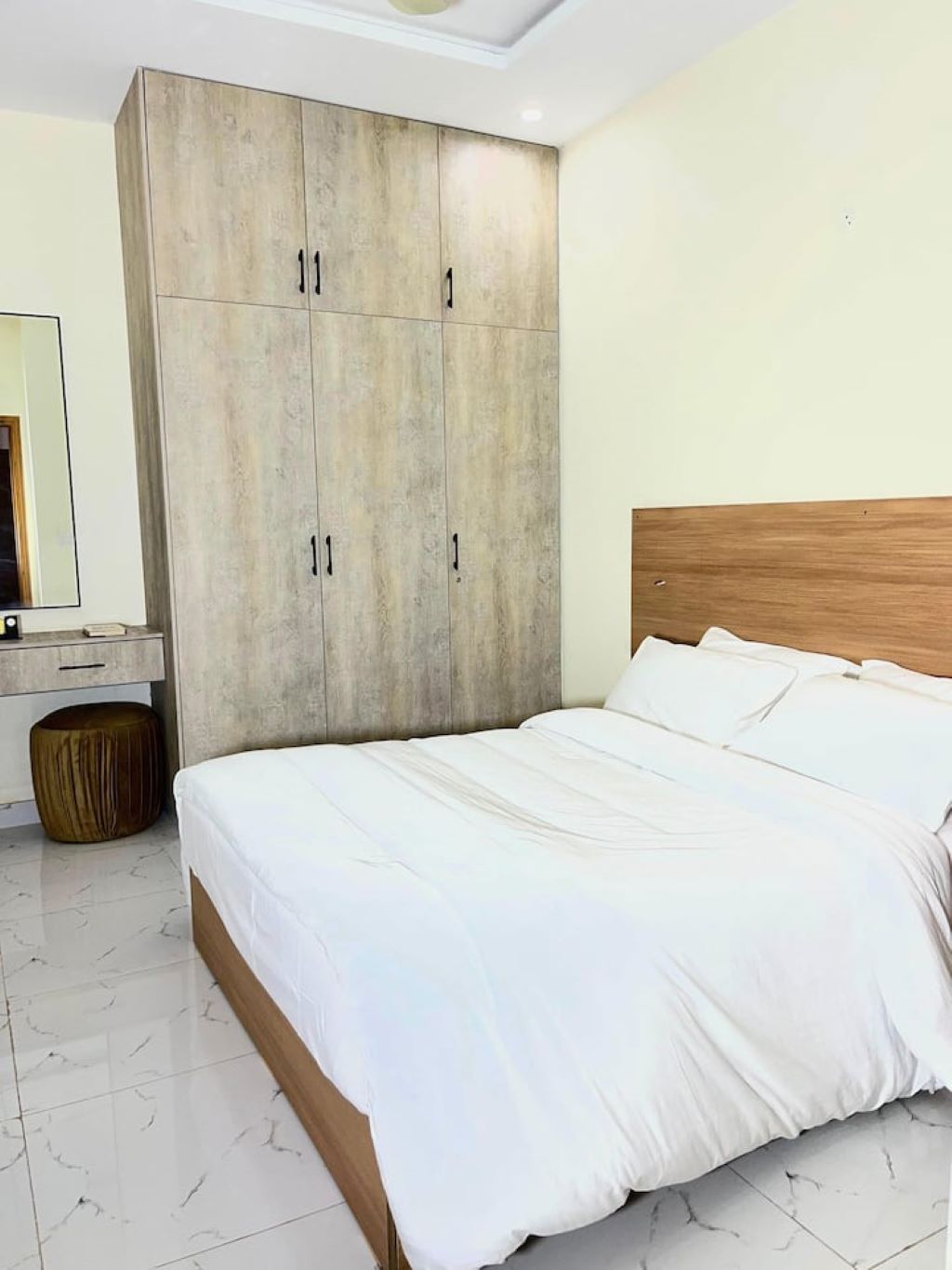 A bedroom with a bed, a wardrobe, a round ottoman, and a mirror.