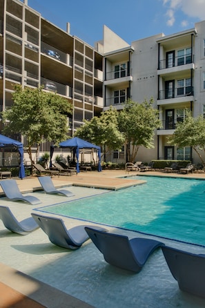 A pool area with lounge chairs and umbrellas, surrounded by multi-story buildings with balconies. - The Bentley Collection - The Jewel (Dallas)