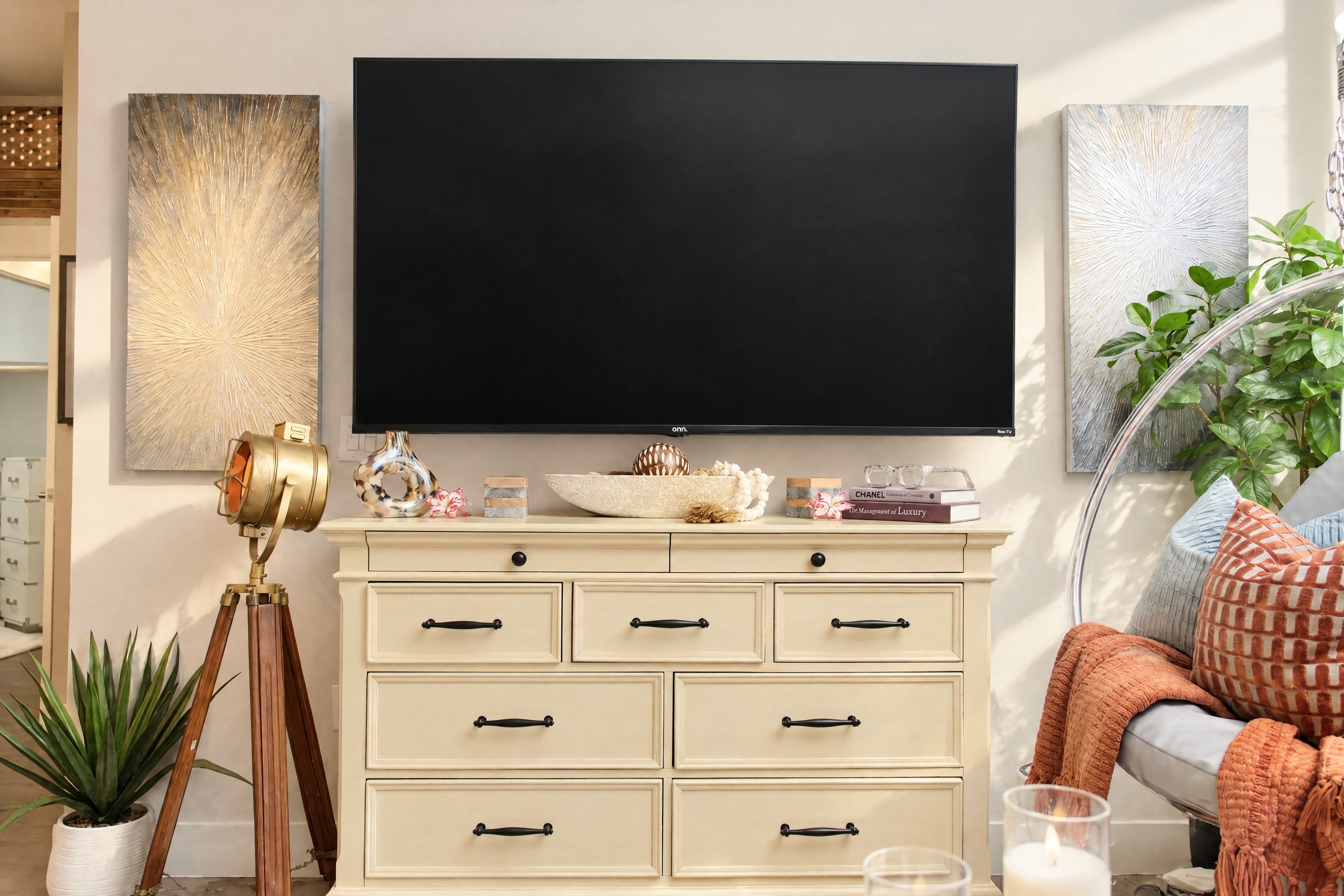 A living room with a large flat-screen TV mounted on the wall, a cream-colored dresser with multiple drawers, a vintage telescope, and a potted plant.