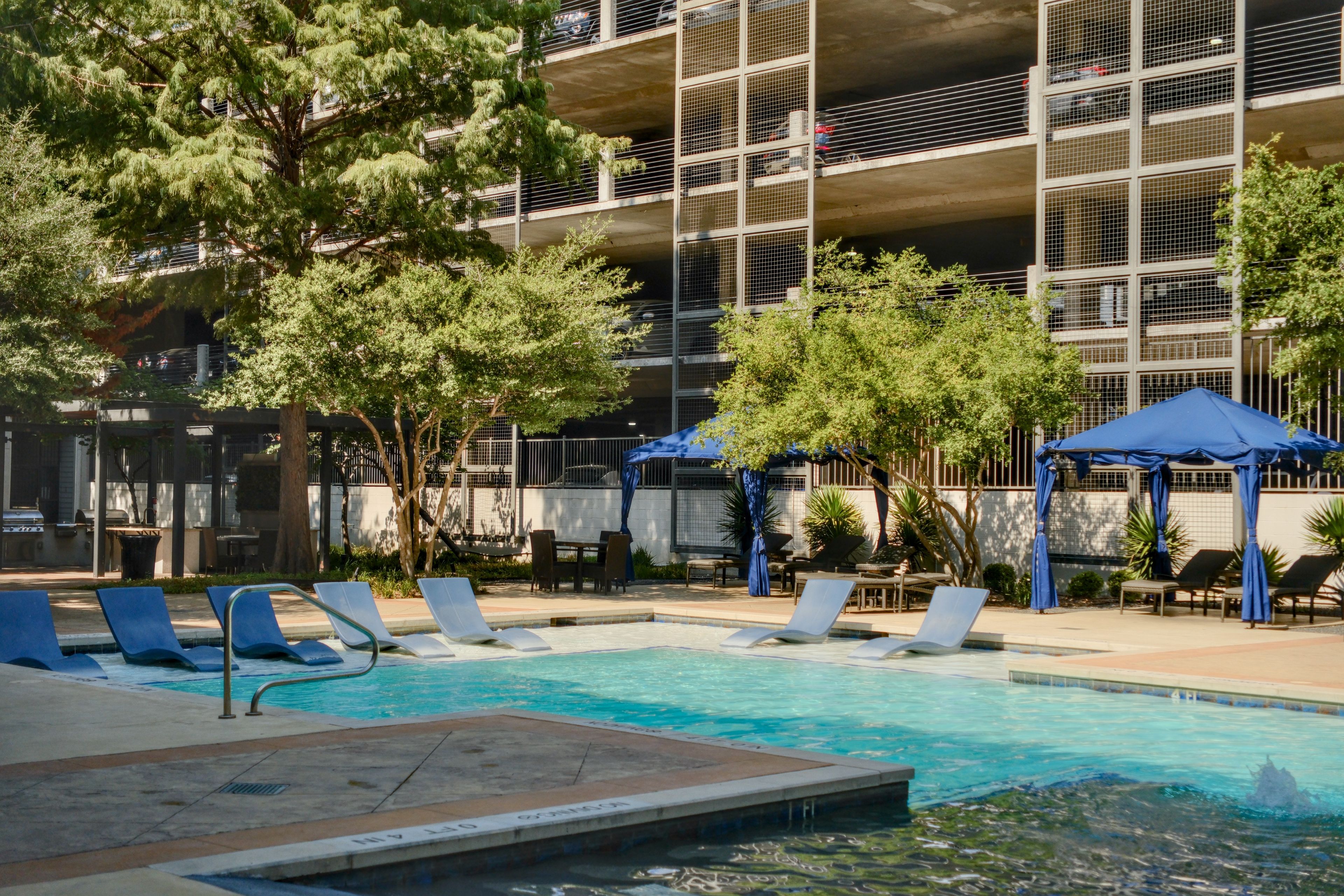 A swimming pool with lounge chairs and umbrellas, surrounded by trees and a building with a parking garage.