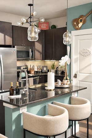 A modern kitchen with a central island, bar stools, stainless steel appliances, and decorative elements. - The Bentley Collection - The Jewel (Dallas)