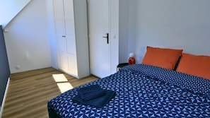 Ein Schlafzimmer mit einem Bett, einem orangefarbenen Kissen, einer blauen, gemusterten Tagesdecke, einer weißen Tür und einem Holzboden.