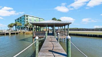 A wooden pier leading to a gazebo with a view of a building and palm trees.