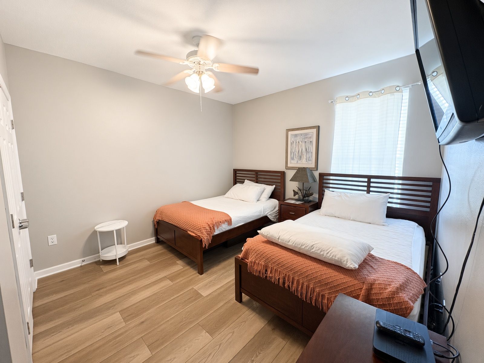 A hotel room with two single beds, a ceiling fan, a small bedside table with a lamp, and a television.