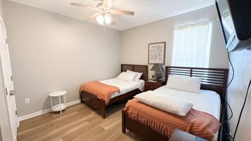 A hotel room with two single beds, a ceiling fan, a small bedside table with a lamp, and a television.
