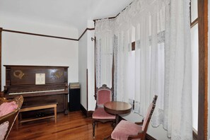 Une pièce avec un piano en bois, un fauteuil recouvert d’un tissu rouge, une petite table ronde et des rideaux en dentelle.