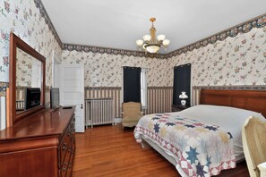 Une chambre à coucher avec un papier peint à motifs floraux, une commode en bois, un lit recouvert d’une courtepointe, une chaise, un radiateur et un lustre.