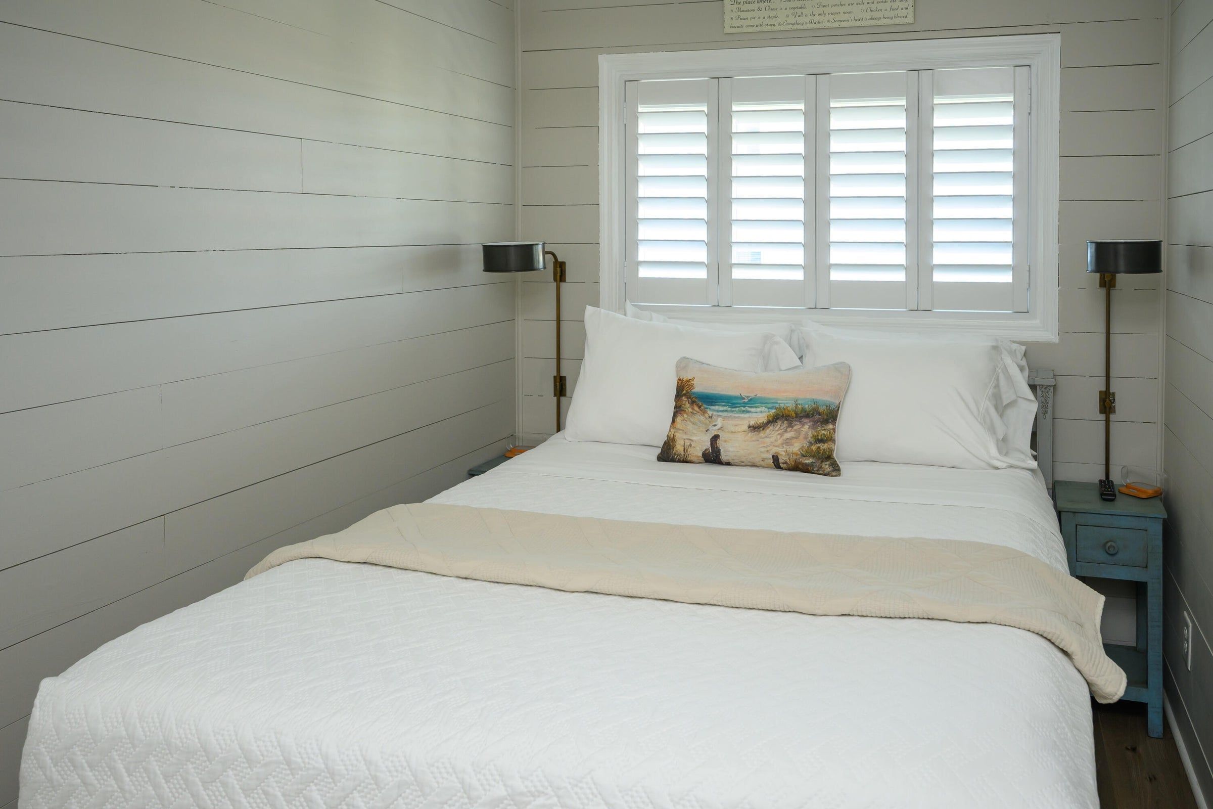 A bedroom with a bed, a window with blinds, a nightstand with a lamp, and a wall with a sign.