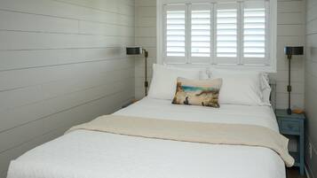 A bedroom with a bed, a window with blinds, a nightstand with a lamp, and a wall with a sign.