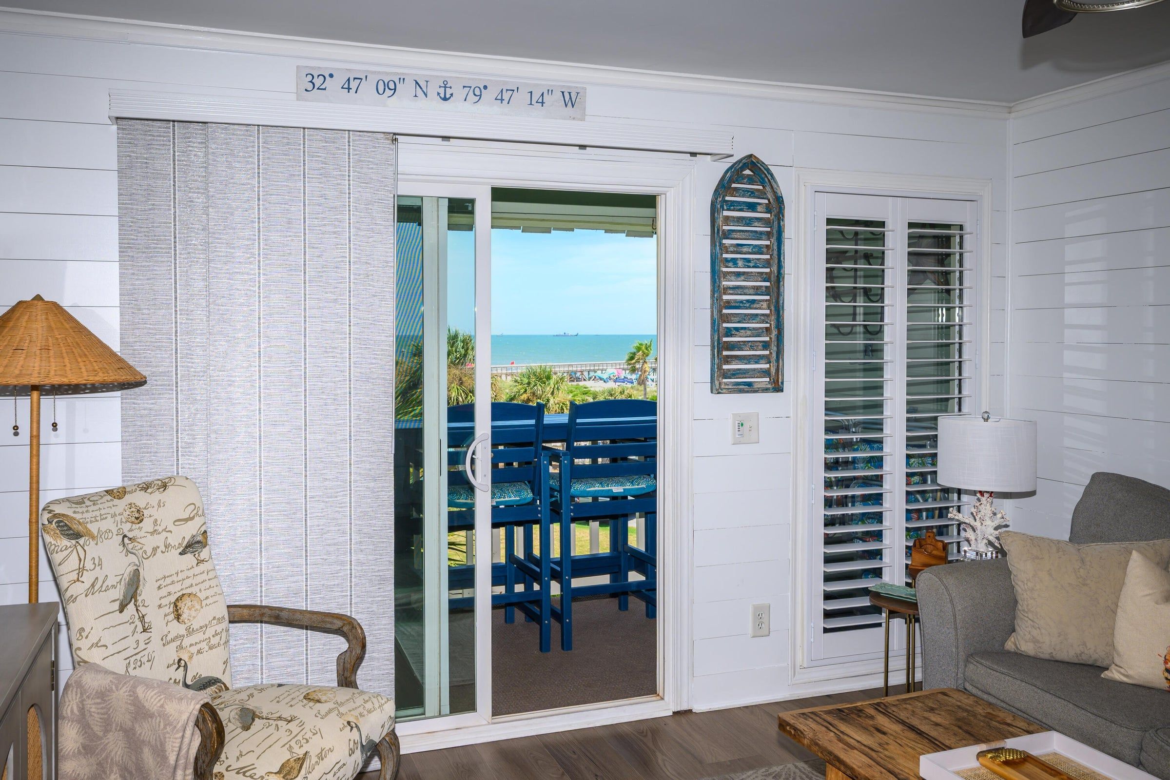 A room with a view of the beach, a door with a nautical-themed decoration, a chair with a bird pattern, and a lamp.