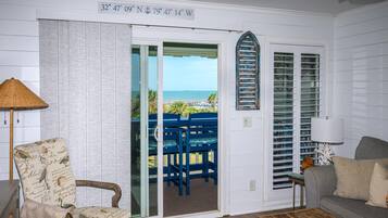 A room with a view of the beach, a door with a nautical-themed decoration, a chair with a bird pattern, and a lamp.