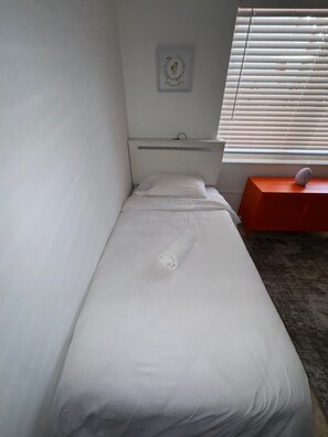 A single bed with a headboard, a small bedside table with a decorative item, and a window with blinds.