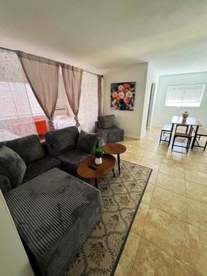 A living room with a sectional sofa, a small coffee table, a dining table with chairs, and a painting on the wall.