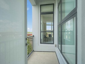 A balcony with a glass door, a concrete floor, and a view of a building with a table and chairs.