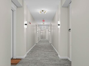 A hotel hallway with a grey carpet, white walls, and a ceiling light fixture.