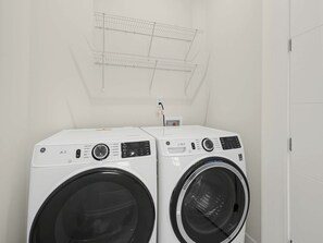 Two front-loading washing machines in a laundry room with a metal shelf above and a power outlet on the wall. - Brand-New Luxury Furnished Penthouse in NoDa, MidTerm Rental (Charlotte)