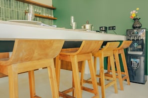 Une cuisine avec un plan de travail blanc, des tabourets de bar en bois, une machine à café et un réfrigérateur.