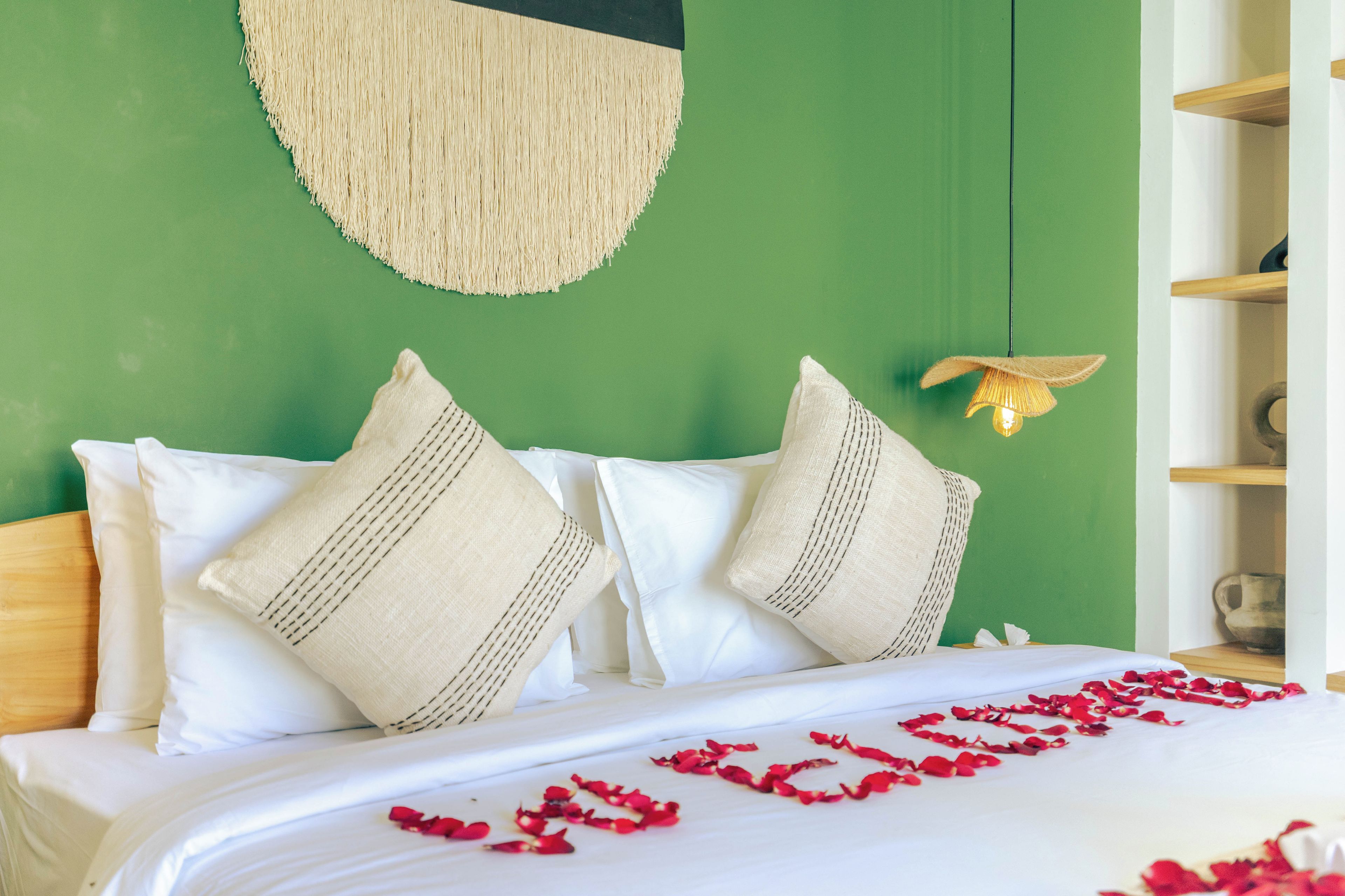A bed with white linens and two decorative pillows, a woven wall hanging above, and a small shelf with a decorative item.