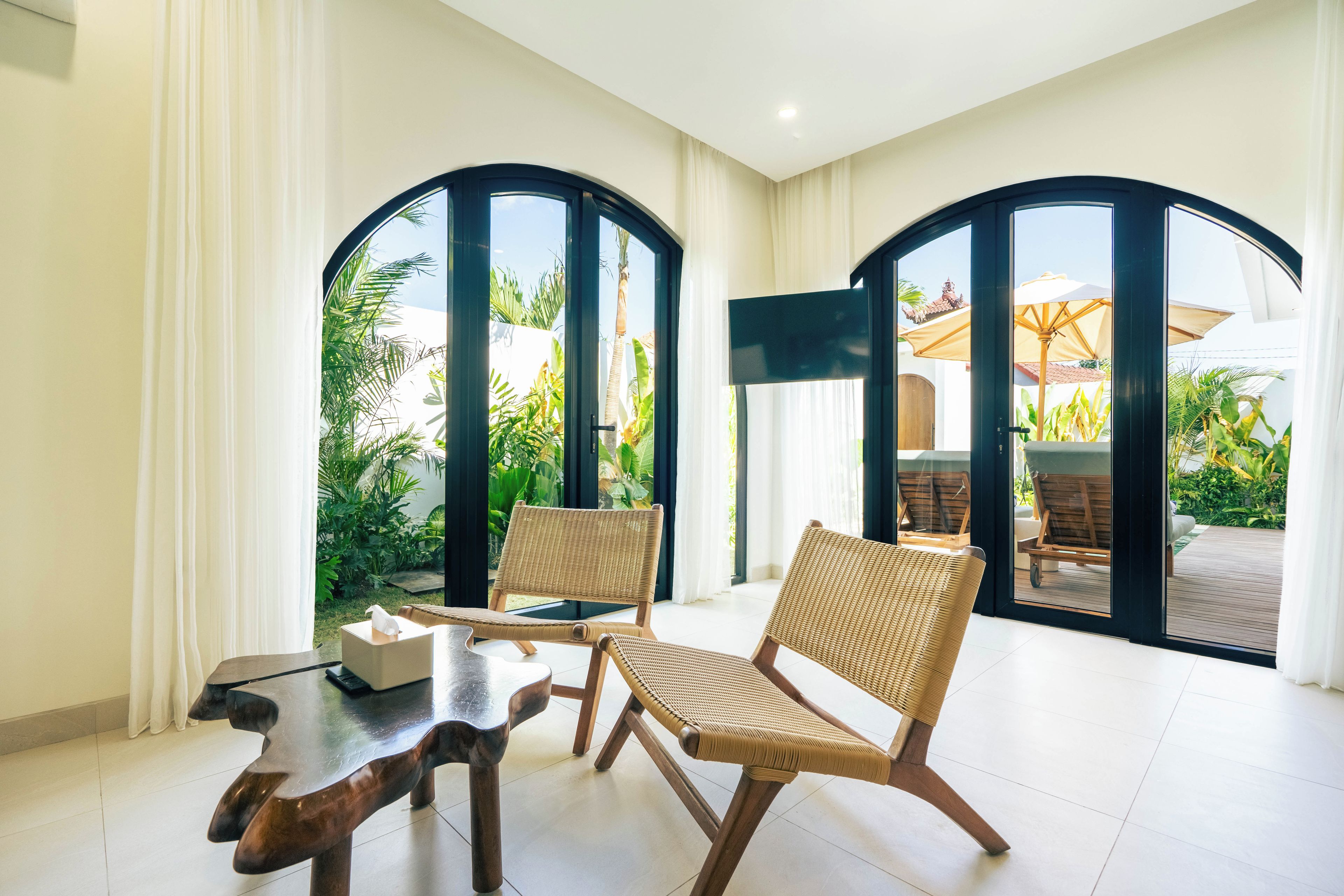 Una habitación con dos sillones de mimbre, una mesa de madera y un ventanal con vistas a un jardín.