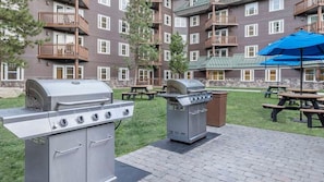 Un espace extérieur aménagé en patio, avec un barbecue et des tables de pique-nique, le tout entouré d’un bâtiment doté de balcons.