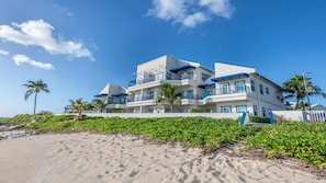 A multi-story beachfront hotel with balconies, palm trees, and a sandy beach in the foreground.